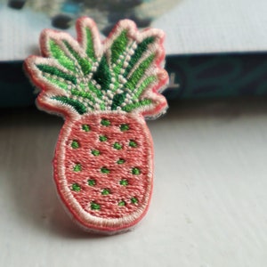 May include: An embroidered pineapple patch in pink and green with white stitching around the edges.