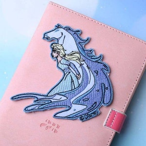 May include: A blue and white embroidered patch featuring a woman riding a horse. The patch is on a pink background and has the text "Disney" on it.
