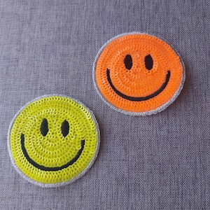 May include: Two smiley face patches, one yellow and one orange, made of sequins. The patches are on a gray fabric background.