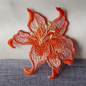 May include: Embroidered orange flower patch with delicate details. The flower has a layered design with a lighter pink hue on the petals' edges. The centre features yellow and green accents, creating a vibrant floral design.