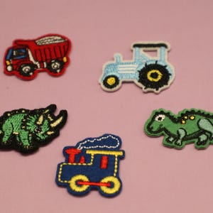 May include: Five embroidered patches featuring a red dump truck, a light blue tractor, a green triceratops, a blue train, and a green dinosaur. Each patch has a black border and is set against a pink background.