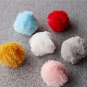 May include: A collection of six fluffy pom-poms in various colors: light blue, red, white, yellow, gray, and pink. These soft, round decorative elements are arranged on a gray fabric surface.
