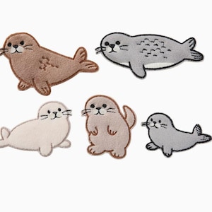 May include: A set of five embroidered patches featuring various seal designs. The collection includes two brown seals, two grey seals with black detailing, and one cream-coloured seal, all depicted in different poses with stitched facial features.