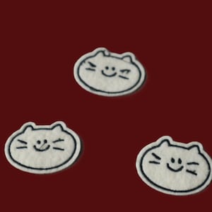 Peut inclure: Trois écussons en feutrine blanche en forme de têtes de chat avec des contours noirs brodés. Chaque écusson présente un visage simple cousu avec deux petites oreilles, trois moustaches de chaque côté et une ligne courbe pour la bouche.