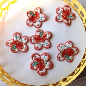 May include: Six pink embroidered flower patches with white and green accents. The patches are on a yellow background.