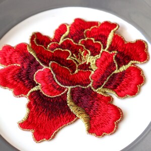 May include: An embroidered flower patch in red and gold. The flower is a peony with a detailed design and a gold outline.