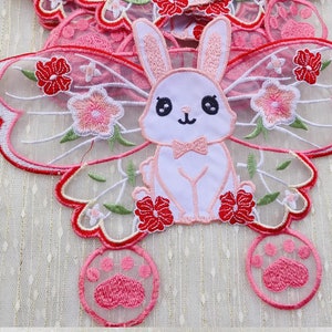 May include: Embroidered patches featuring a white bunny with a pink bow tie, surrounded by pink and red flowers and butterfly-shaped wings. The patches also include pink paw prints. The design is on a sheer, light pink background.