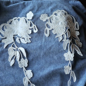 May include: Two white lace appliques with floral designs on a blue denim background.