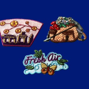 Peut inclure: Trois écussons brodés : un écusson en forme d'éventail avec des fleurs jaunes et un bâtiment violet, un écusson en forme de panier tressé marron avec des fleurs rouges et blanches, et un écusson avec le texte Fresh Air, des feuilles vertes et un papillon jaune et orange.