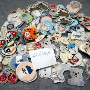 May include: A selection of embroidered patches in diverse shapes and colours. Designs feature animals such as bears, tigers, and dogs, alongside other whimsical motifs. The patches are crafted with colourful threads and have a sewn appearance. A handwritten note reads "sew100A".