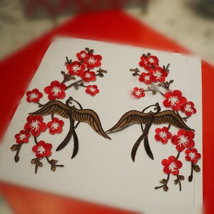 May include: Two embroidered patches featuring brown birds with white bellies and red blossoms. The birds are flying with their wings spread. The blossoms are arranged in a branch-like pattern.