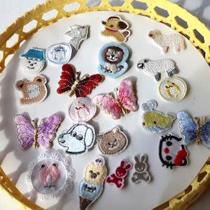 May include: A collection of colourful embroidered iron-on patches featuring various animal designs, including butterflies, a lion, a sheep, a dog, a cat, and a frog. The patches are arranged on a white plate with a yellow wicker border.