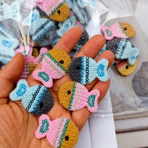 May include: A collection of small, crocheted fish-shaped appliques in various color combinations including pink, yellow, light blue, and dark gray. Each piece features a distinct cross-stitch pattern detail and a small circular opening for an eye.