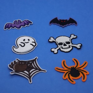 May include: A set of eight Halloween-themed iron-on patches. The patches feature a spider, a spider web, a ghost, a skull and crossbones, and two bats. The patches are in various colours, including black, white, orange, purple, and red.