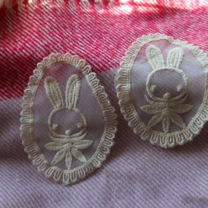 May include: Two white lace bunny appliques against a pink and white striped background. The appliques are oval shaped and have a delicate lace trim.