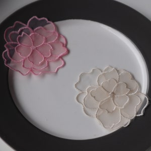 May include: Two delicate lace flower appliques, one pink and one cream, on a white plate with a black rim.