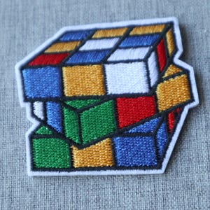 May include: Embroidered patch featuring a colorful Rubik's Cube design. The patch showcases the cube's classic arrangement of colored squares: red, blue, green, yellow, white, and orange. The patch has a white border and is likely for application on clothing or accessories.