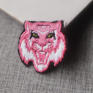 May include: An embroidered patch featuring a stylized tiger head in shades of pink and white with yellow eyes. The tiger is depicted with an open mouth showing teeth, set against a black background border, creating a detailed textile accessory.