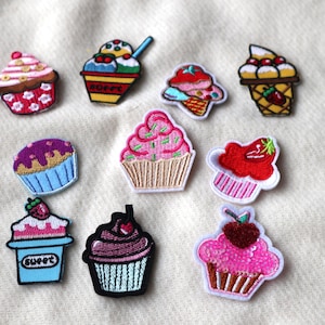 May include: A selection of embroidered cupcake patches in various colours. Each patch showcases a different cupcake design, with details like frosting, sprinkles, and cherries. Some patches include the word "sweet". The patches are displayed on a light-coloured fabric.