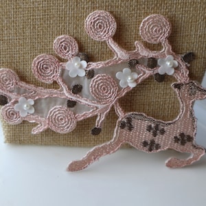 May include: A pink and brown embroidered deer applique with white pearl-like embellishments. The deer is running and has a floral design on its back.