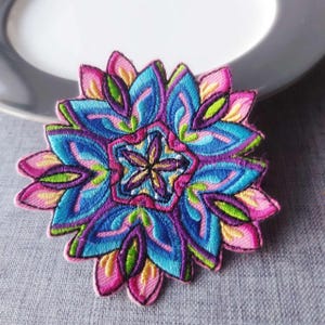 May include: Embroidered patch featuring a vibrant floral design. The patch showcases a symmetrical pattern with shades of blue, pink, purple, yellow, and green. The intricate stitching creates a detailed, textured appearance, perfect for adding flair to clothing or accessories.