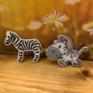 May include: Two embroidered zebra patches. One shows a standing zebra with black and white stripes. The other depicts a cartoon zebra in a running pose, with grey and white stripes and a grey head.