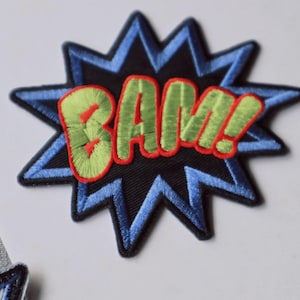 May include: An embroidered patch shaped like a comic book speech bubble with a black centre and blue starburst border. The word BAM! is stitched in light green thread with a bold red outline, creating a vibrant, multi-coloured graphic design.