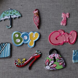 May include: A selection of embroidered patches in diverse shapes and colours. Features an umbrella, dress, bow, the word "BOY", the word "GIRL", a kimono, a high heel, a purse, and denim shorts. Patches are on a grey fabric background.