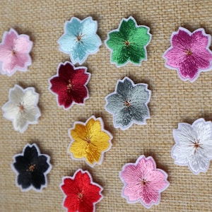 May include: Assortment of 12 embroidered iron-on patches in various colors, each featuring a five-petal flower design. The flowers are in shades of pink, red, white, blue, green, yellow, and black.