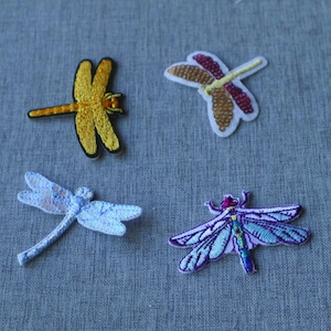 Peut inclure: Quatre écussons brodés en forme de libellule de couleurs et styles différents. Un est jaune uni, un présente des sequins marron et rouges, un est bleu clair avec une finition texturée, et un est violet et bleu avec des ailes finement brodées.