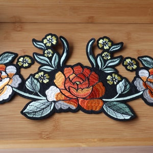 May include: An embroidered patch featuring three orange and white flowers, green leaves, and yellow flower buds on a black background.