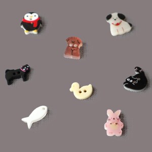 May include: A collection of decorative buttons in various animal shapes, including a penguin, dog, cat, duck, fish, and rabbit. The buttons are colorful and detailed, perfect for sewing or craft projects.