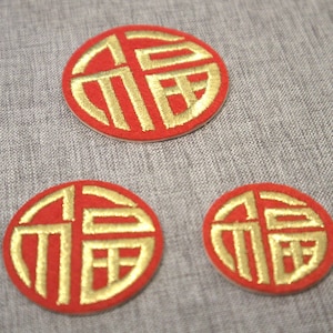 May include: Three red, circular embroidered patches with gold Chinese characters. The patches are in different sizes and are arranged on a grey fabric surface. The gold embroidery is detailed against the red background.