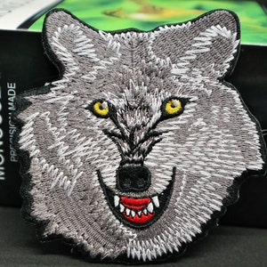 May include: Iron-on patch featuring an embroidered grey wolf's face with sharp teeth and yellow eyes. The wolf's mouth is open, revealing red teeth.