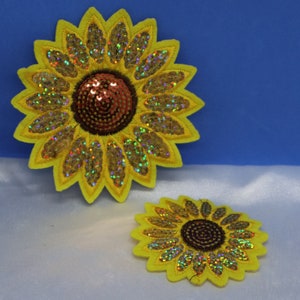 May include: Two yellow felt sunflower patches with iridescent sequins and red centers. The larger sunflower is about 5 inches in diameter and the smaller sunflower is about 3 inches in diameter.