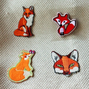 May include: Four embroidered fox patches in different designs. One fox is sitting, another is curled up, a third has a flower, and the last is a fox face. The patches are orange, white, and black with green detailing.