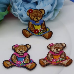 Könnte beinhalten: Drei gestickte Bügelbilder mit braunen Teddybären, die bunte Kleider tragen. Die Bären sitzen mit gekreuzten Armen da und halten Blumen in den Pfoten.