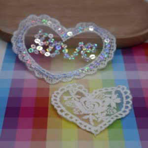 Peut inclure: Deux appliques en dentelle de forme de cœur blanches. La plus grande applique est décorée de paillettes irisées qui épellent "love".