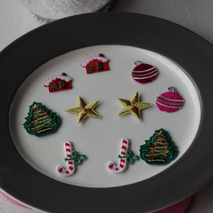 May include: Embroidered Christmas patches featuring red houses with white snow, gold stars, green Christmas trees, red and white candy canes, and pink and red baubles. The patches are arranged on a white plate with a grey rim.