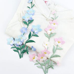 Peut inclure: Trois patchs de fleurs brodés en bleu, rose et blanc. Les patchs présentent des motifs floraux délicats avec des tiges et des feuilles vertes.