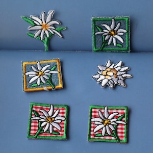 May include: Six embroidered iron-on patches featuring white edelweiss flowers with green stems and yellow centres. The patches are on a blue background. Some patches have a blue denim background, others have a red and white checked background.