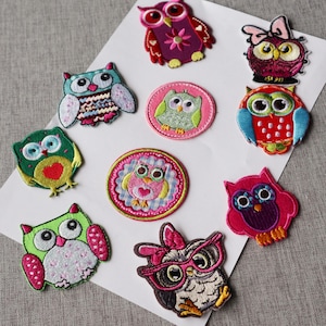 Peut inclure: Plusieurs écussons brodés en forme de hiboux, aux couleurs vives comme le rose, le vert, le bleu et le violet. Chaque patch présente un design de hibou unique, avec différents motifs et accessoires, tels que des nœuds et des lunettes. Les écussons sont disposés sur une surface blanche.