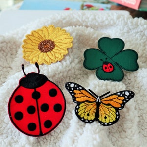 May include: Four embroidered iron-on patches: a yellow sunflower, a green four-leaf clover with a red ladybug, a red ladybug with black spots, and a yellow and orange butterfly.