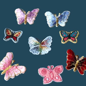 May include: A collection of embroidered butterfly patches in various colors, including pink, purple, blue, and red. Each patch features detailed stitching and gold accents, showcasing a variety of butterfly designs. The patches are displayed against a teal background.