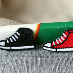 May include: Two embroidered iron-on patches of high-top sneakers. One patch is black with white laces and the other is red with white laces.