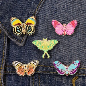 May include: Five butterfly enamel pins in various colours. The pins feature different designs, including a yellow and black butterfly, a pink butterfly, a green moth, an orange and white butterfly, and a blue and purple butterfly. They are on a denim background.