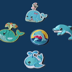 May include: Five embroidered patches featuring whales and a dolphin. The patches are in shades of blue, yellow, and white, set against a dark blue background. The patches are suitable for clothing or accessories.