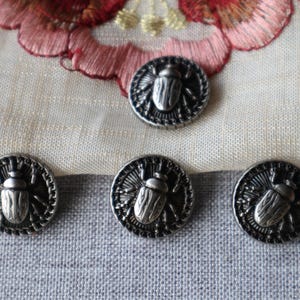 May include: Four antique silver-coloured buttons with a detailed beetle design. The buttons are round with a textured border. The background includes a light-coloured fabric and a portion of an embroidered floral design.