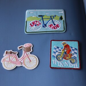 May include: Three embroidered iron-on patches. One features a blue bicycle with the words "Let's Ride" on the front tyre. Another features a pink bicycle with a basket of flowers. The last patch features a blue and red dirt bike with the word "Winner" on the bottom.