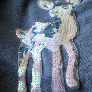 May include: A sequined deer applique in pastel colors of pink, blue, and silver is sewn onto a denim fabric. The deer is outlined with a sheer, light-colored mesh. The sequins create a shimmering effect, adding a decorative touch to the denim.